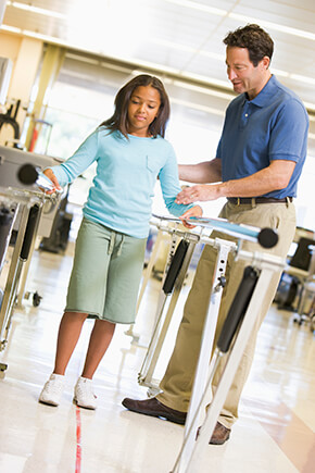 Child walking between parallel bars during a physical therapy session while an adult provider stands beside and offers support in a clinic setting.