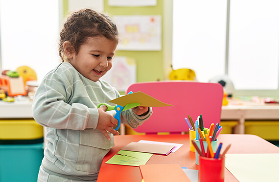 A young girl is standing at her school desk while cutting paper