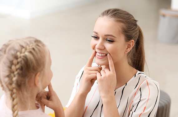 A speech therapist is smiling and pointing to the corners of her mouth while the little girl in front of her copies the same expression